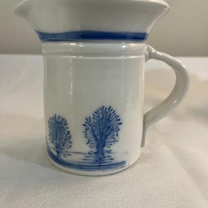 May include: White ceramic pitcher with a blue band around the top and a blue design of trees and water on the bottom.