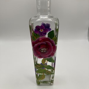 May include: A clear glass bottle with a square base and a narrow neck, featuring a hand-painted design of a large, vibrant magenta rose and smaller purple flowers with green leaves and stems wrapped around the front surface of the glass.