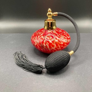 May include: A round glass perfume bottle with a red and white mottled pattern and gold metallic accents. The bottle features a gold-tone atomizer pump connected to a black fabric-covered hose and a black bulb with a long black tassel attached.