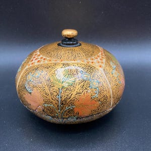 May include: A round, decorative box with a lid. The box is primarily gold with intricate floral and geometric patterns in orange, green, and blue. The lid has a small, gold knob.