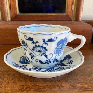 May include: A white porcelain teacup and saucer set featuring a scalloped edge and a blue floral pattern. The design includes intricate blue botanical motifs, swirling vines, and stylized blossoms painted across the exterior of the cup and the saucer.