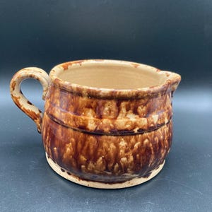 May include: A brown and tan ceramic pitcher with a handle and spout. The pitcher has a rustic, glazed finish with dark brown streaks and bands around the body. The interior is a lighter tan color.