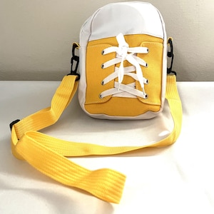 May include: A small white and yellow canvas crossbody bag with a white lace-up design. The bag has a yellow adjustable strap.