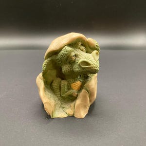 May include: A small, detailed figurine of a baby dinosaur hatching from an egg. The dinosaur is green with textured skin, and the eggshell is tan. The figurine is on a dark surface.
