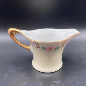 May include: Cream-colored porcelain creamer with a gold handle and rim. The creamer features a pink and gold floral design with a delicate pink flower and green leaves. A classic piece for serving cream or milk.