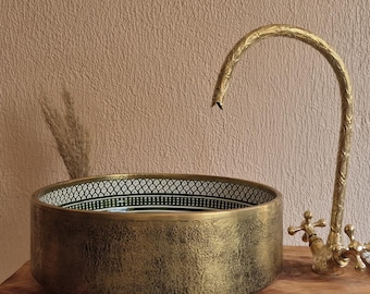 May include: A gold-toned, round bathroom sink with a patterned rim and a matching faucet. The sink sits on a wooden base. The faucet has a curved spout and decorative handles. The sink's interior is green.