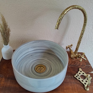 May include: A round, marble-effect sink with a brass faucet and drain. The faucet has an ornate design. A small white vase with dried grass sits to the left of the sink. The sink is on a wooden surface.