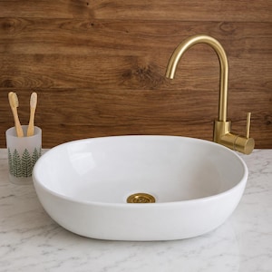 May include: A white oval ceramic vessel sink with a gold-toned metal tap and matching drain. Next to the sink is a frosted glass toothbrush holder containing two wooden toothbrushes with a green pine tree pattern printed on the glass.