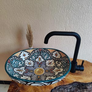 May include: An oval ceramic basin with a floral design in teal, white, red, and gold. It has a black tap and rests on a natural wood base. The basin's pattern includes detailed elements and a decorative drain.