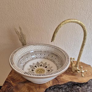 May include: A decorative ceramic sink with a detailed floral pattern in shades of brown and cream. The sink is paired with a curved gold tap. The sink is placed on a wooden surface.