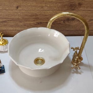Op de afbeelding: Cr&egrave;mekleurige keramische wastafel met een geschulpte rand en een goudkleurige kraan. De kraan heeft een gebogen uitloop en kruisvormige handgrepen. Een kleine goudkleurige afdekking is zichtbaar. De wastafel staat op een wit oppervlak.