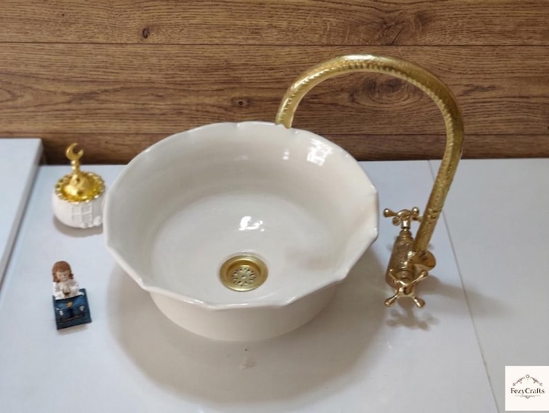 Op de afbeelding: Cr&egrave;mekleurige keramische wastafel met een geschulpte rand en een goudkleurige kraan. De kraan heeft een gebogen uitloop en kruisvormige handgrepen. Een kleine goudkleurige afdekking is zichtbaar. De wastafel staat op een wit oppervlak.