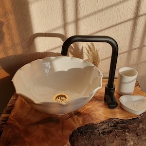 May include: A cream-colored, scalloped-edge ceramic sink with a gold drain cover. A black faucet is installed. A matching ceramic cup and soap dish sit beside the sink. The sink is on a wooden surface.