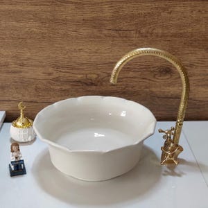 Op de afbeelding: Een cr&egrave;mekleurige keramische wastafel met geschulpte randen en een goudkleurige kraan. De kraan heeft een sierlijk ontwerp. Decoratieve items zijn bij de wastafel geplaatst. De achtergrond is een houten muur.