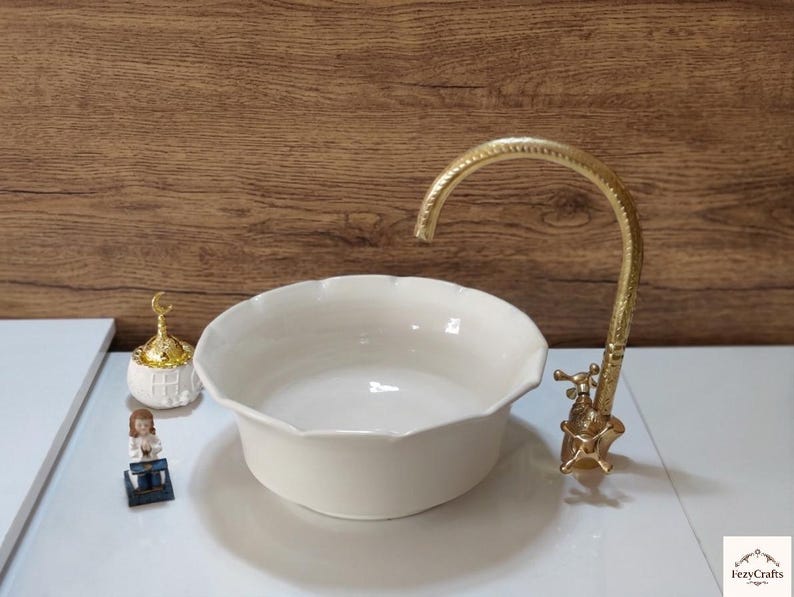 Op de afbeelding: Een cr&egrave;mekleurige keramische wastafel met geschulpte randen en een goudkleurige kraan. De kraan heeft een sierlijk ontwerp. Decoratieve items zijn bij de wastafel geplaatst. De achtergrond is een houten muur.