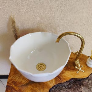 May include: A white ceramic vessel sink with a scalloped rim and a brass faucet. The sink is set on a natural wood base. A small decorative lamp sits to the right of the faucet.