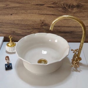 Op de afbeelding: Een cr&egrave;mekleurige keramische wastafel met geschulpte randen en een goudkleurige kraan. De wastafel staat op een wit aanrechtblad. Decoratieve items zijn een kleine goudkleurige container en een beeldje.
