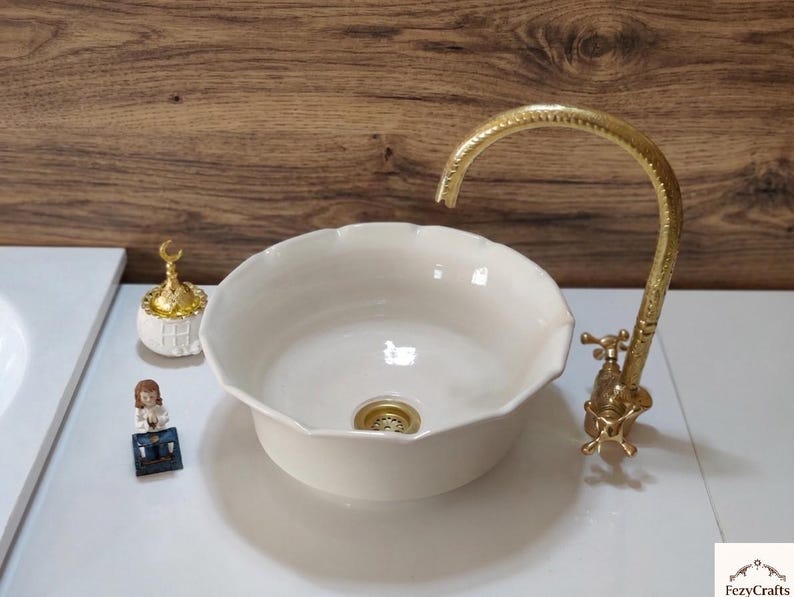 Op de afbeelding: Een cr&egrave;mekleurige keramische wastafel met geschulpte randen en een goudkleurige kraan. De wastafel staat op een wit aanrechtblad. Decoratieve items zijn een kleine goudkleurige container en een beeldje.