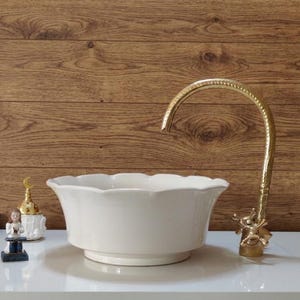 Op de afbeelding: Een witte keramische wastafel met een geschulpte rand, gecombineerd met een goudkleurige kraan. De kraan heeft een sierlijk ontwerp. De wastafel staat op een wit oppervlak, met een houten achtergrond. Decoratieve items zijn links van de wastafel zichtbaar.