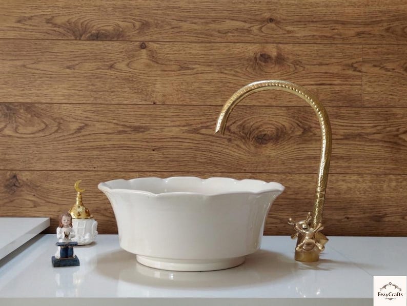Op de afbeelding: Een witte keramische wastafel met een geschulpte rand, gecombineerd met een goudkleurige kraan. De kraan heeft een sierlijk ontwerp. De wastafel staat op een wit oppervlak, met een houten achtergrond. Decoratieve items zijn links van de wastafel zichtbaar.