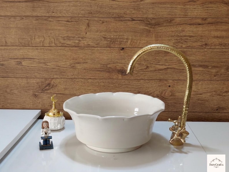 Op de afbeelding: Een cr&egrave;mekleurige keramische wastafel met geschulpte randen en een goudkleurige kraan. De kraan heeft een sierlijk ontwerp. Een klein decoratief item en een beeldje staan op het aanrecht. De achtergrond is een houten wand.