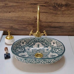 May include: An oval ceramic vessel sink featuring a dark green and white intricate floral pattern with a gold-toned drain. A tall, textured gold-toned faucet stands behind the basin. A small white and gold decorative object sits to the left.