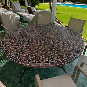 Op de afbeelding: Een ronde eettafel voor buiten met een mozaïek tafelblad bestaande uit kleine, vierkante tegels in bruine, zwarte en beige tinten in een geometrisch kubuspatroon, ondersteund door een zwart metalen frame met vier poten.