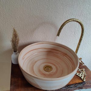 May include: A ceramic vessel sink with a cream and brown swirl pattern. The sink has a brass drain and is paired with a curved brass faucet. A small white vase with dried flowers sits to the left of the sink. The sink is on a wooden surface.