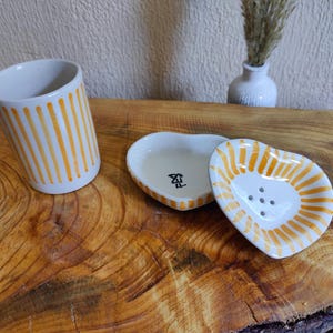 Puede incluir: Una taza de cerámica blanca y dos platos en forma de corazón. La taza y los platos están decorados con rayas verticales naranjas. Un plato es un jabonera con agujeros de drenaje.