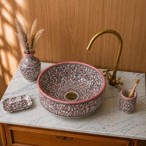 Puede incluir: Un lavabo de cerámica redondo con un patrón floral rosa y negro, acompañado de una jabonera, un portacepillos y un jarrón a juego. Un grifo de cuello de cisne en tono latón se sitúa detrás del lavabo sobre una encimera de mármol blanco.