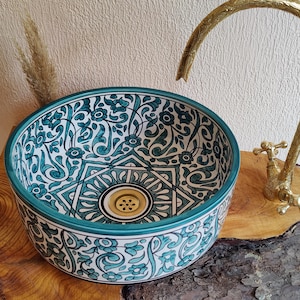 Può includere: Un lavabo rotondo in ceramica con un motivo floreale turchese e bianco. Il lavabo ha uno scarico dorato ed è abbinato a un rubinetto dorato. Il lavabo è posto su una superficie di legno.