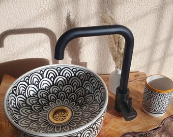 May include: A white ceramic sink with a black and white geometric pattern. The sink has a gold drain and is sitting on a wooden countertop. A black faucet is mounted to the countertop.