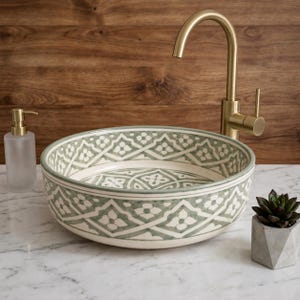 Puede incluir: Un lavabo de cerámica redondo con un patrón floral geométrico en verde salvia y blanco. El lavabo se combina con un grifo alto de oro cepillado. A la izquierda hay un dispensador de jabón de vidrio y a la derecha una pequeña suculenta.