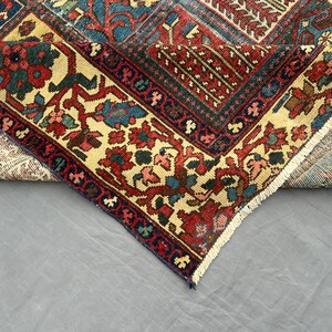 May include: A folded area rug featuring a complex geometric and floral pattern in shades of deep red, navy blue, and cream. The rug displays a dense arrangement of intricate motifs, a wide decorative border, and a short white fringe along the edge.