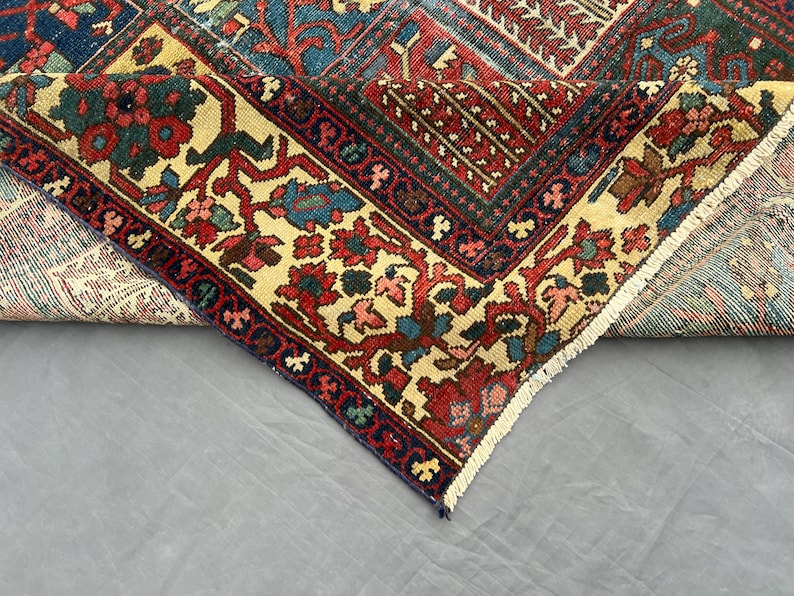 May include: A folded area rug featuring a complex geometric and floral pattern in shades of deep red, navy blue, and cream. The rug displays a dense arrangement of intricate motifs, a wide decorative border, and a short white fringe along the edge.