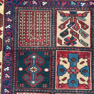 May include: A woven rug featuring a grid of rectangular panels with intricate geometric and floral patterns in shades of deep red, navy blue, and cream. The border displays a repeating floral motif on a cream background with blue accents.