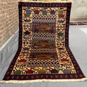 Op de afbeelding: Een rechthoekig vloerkleed met een centraal donkerblauw paneel met herhalende geometrische motieven in bruin, rood en groen. De rand heeft een crèmekleurige achtergrond met gestileerde vaatpatronen en donkere franjes aan beide uiteinden.