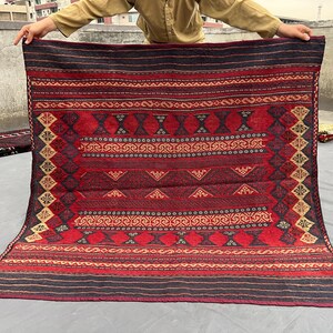 May include: A rectangular woven textile featuring a deep red base color with horizontal bands of black and cream geometric patterns. The design includes repeating diamond shapes along the vertical edges and intricate tribal-inspired motifs throughout.