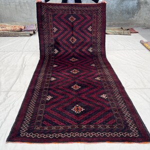 Pode incluir: Um tapete longo e retangular com um padrão geométrico de diamantes em tons de bordô e preto. O tapete tem uma borda detalhada e pequenos elementos decorativos no centro. O tapete é exibido em uma superfície branca.