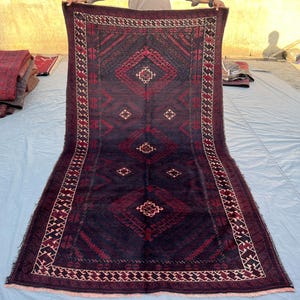 Pode incluir: Um tapete retangular, bordô escuro, com um padrão geométrico. O tapete apresenta desenhos de diamantes e ziguezagues em vermelho e creme, sobre um fundo azul escuro. A borda tem um padrão repetitivo de formas vermelhas e creme. O tapete é exibido ao ar livre.