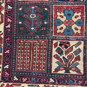 May include: A woven rug featuring a grid pattern of square panels. Each panel contains distinct geometric and floral motifs in shades of deep red, navy blue, cream, and teal. The border displays intricate repeating patterns in matching colours.