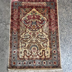 May include: An ornate, rectangular rug with a cream-colored central field, framed by a border of burgundy, blue, and beige. The rug features intricate floral and geometric patterns, with a fringe along the edges. The rug measures approximately 36 inches long and 24 inches wide.