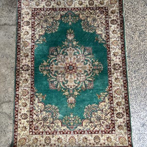 May include: A rectangular, ornate rug with a central teal field and a detailed floral pattern. The border is cream with red accents. The rug has a fringed edge and a traditional design.