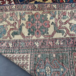 May include: A woven rug featuring a cream-coloured border with repeating floral motifs in shades of pink, blue, and dark green. The main body of the rug displays a geometric pattern in deep blue, red, and cream, with a corner folded over to reveal.