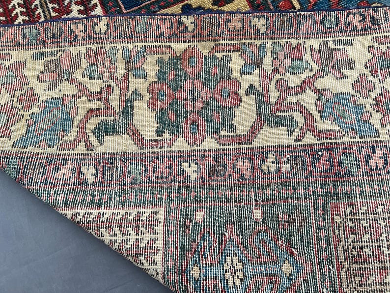 May include: A woven rug featuring a cream-coloured border with repeating floral motifs in shades of pink, blue, and dark green. The main body of the rug displays a geometric pattern in deep blue, red, and cream, with a corner folded over to reveal.