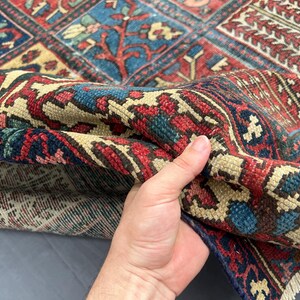 May include: A woven area rug featuring a complex geometric and floral pattern in shades of deep red, navy blue, and cream. The rug displays a dense pile construction with visible knotted texture and a fringed edge along the bottom border.