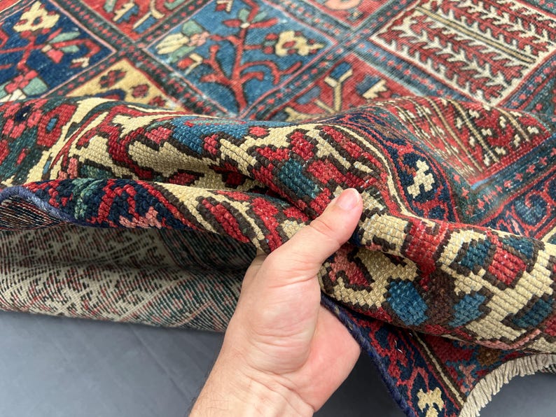 May include: A woven area rug featuring a complex geometric and floral pattern in shades of deep red, navy blue, and cream. The rug displays a dense pile construction with visible knotted texture and a fringed edge along the bottom border.