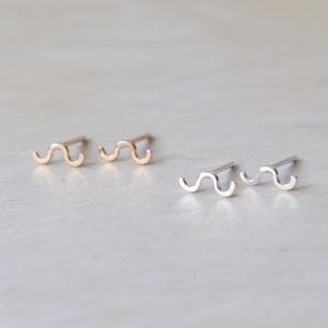 以下が含まれることがあります： 波型のスタッドピアス2組。1組はゴールドトーン、もう1組はシルバートーンです。ピアスは小さく、ミニマルで、シンプルで湾曲したデザインです。明るい色の表面に表示されています。