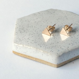 May include: A pair of gold triangle stud earrings on a white speckled tile background.