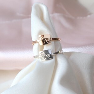 May include: Two silver rings with a square design featuring a sun and wave motif. The ring on top is rose gold.